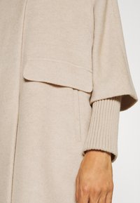 Beige wollen blend jas met een losse pasvorm, voorzien van een omvouwzak en ribgebreide manchetten. Zachte textuur en minimalistisch design.