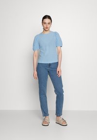 Top texturizado azul claro com mangas puff curtas, combinado com jeans azuis justos e sandálias bege com detalhes decorativos em metal.