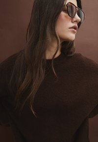Maglione oversized marrone, caratterizzato da una texture morbida, indossato con occhiali da sole scuri e alla moda. Lo sfondo presenta una tonalità di marrone simile.
