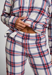Geruite flanellen pyjama set in het wit, met rode en blauwe lijnen. Blouse met knopen, lange mouwen en broek met elastische tailleband. Zachte textuur.