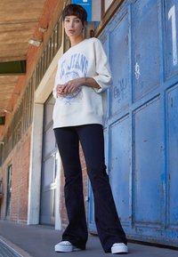 Jeune personne aux cheveux courts portant un sweat-shirt blanc oversized et un pantalon noir évasé, debout à côté d'une porte métallique bleue dans un cadre urbain.