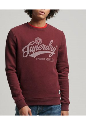 Młody mężczyzna ubrany w bordową bluzę Superdry z czerwoną koszulką pod spodem oraz niebieskie dżinsy, stojący na tle jednolitego, szarego tła.