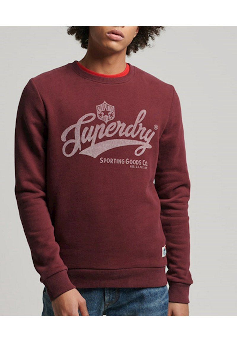 Jeune homme portant un sweat-shirt Superdry bordeaux avec un t-shirt rouge en dessous et un jean bleu, debout devant un fond gris uni.