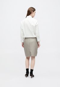 Γυναίκα που στέκεται με την πλάτη προς τα εμάς, φοράει λευκό μακρυμάνικο πουκάμισο, μπεζ φούστα μέχρι τα γόνατα, μαύρες κάλτσες και καφέ παπούτσια με τακούνι.