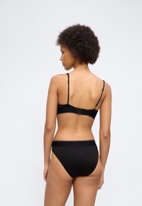 Γυναίκα με κατσαρά μαλλιά που φαίνεται από πίσω, φορώντας μαύρο σουτιέν και μαύρα εσώρουχα, σε απλό ανοιχτό φόντο.