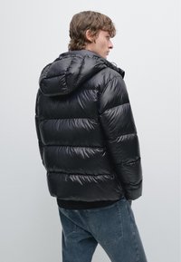 Zwarte pufferjack met een capuchon, voorzien van gestikte delen en een glanzende afwerking. G gedragen over een zwarte trui met blauwe jeans.
