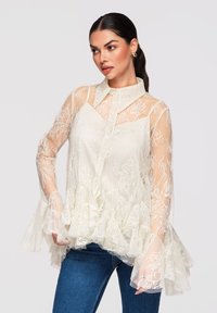 Femme aux cheveux bruns attachés en queue de cheval portant un chemisier en dentelle crème avec des poignets et un ourlet à volants, associé à un jean bleu, regardant sur le côté.