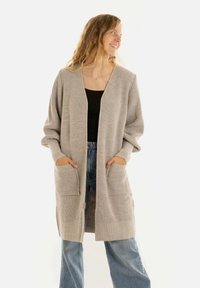 Vrouw die glimlacht en naar rechts kijkt, gekleed in een lange beige gebreide cardigan over een zwarte top en blauwe jeans, met haar handen in de zakken van de cardigan.