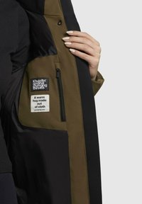 Indvendig udsigt af en jakke med tofarvet design i sort og olivengrøn stof, en lynlåslomme og mærkebånd.