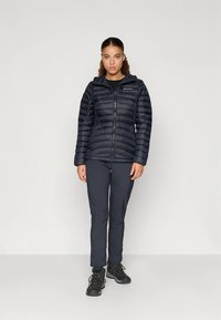 Zwarte donsjack met horizontale stiksels, ritssluiting en capuchon, gecombineerd met donkere broek en zwarte wandelschoenen.