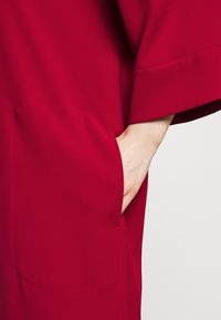 Main placée dans la poche avant d'un vêtement rouge à manches larges, sur un fond clair et uni.