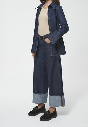 Spijkerjack met een kraag en knopen aan de voorkant, gecombineerd met een broek met wijde pijpen van spijkerstof met een lichterblauwe zoom. Zwarte schoenen met een strikdetail.
