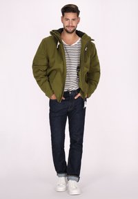 Giacca puffer verde oliva con cappuccio, chiusura zip e due tasche laterali. Abbinata a una maglietta a righe, jeans scuri e sneakers bianche.