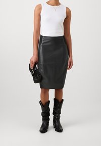 Svart läderpennkjol, figursydd design, slät textur; matchad med en figursydd vit ärmlös topp och svarta cowboystövlar. Håller en liten handväska.