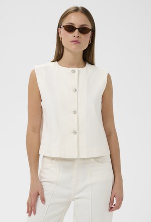 Žena v bezrukávovom bielom zapínanom topu, siahajúcich bielych nohaviciach a úzkych hnedých slnečných okuliaroch, stojaca pred jednoduchým svetlým pozadím.