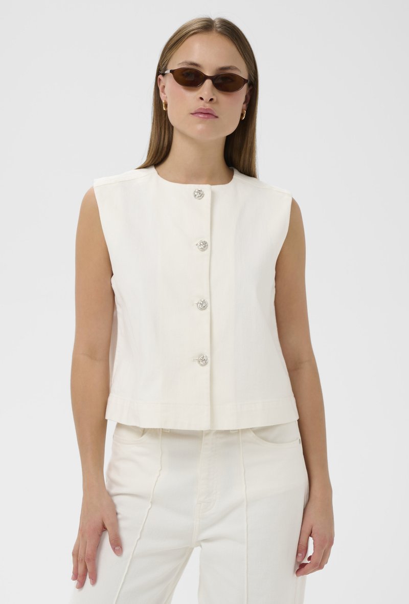 Naine, kes kannab valget varrukateta nööbitavat topi, sobivaid valgeid pükse ja kitsaid pruune päikeseprille, seisab ühevärvilise heleda tausta ees.