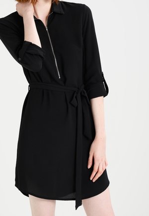 Czarna sukienka z dekoltem na zamek błyskawiczny, długimi rękawami podwiniętymi i materiałowym paskiem w talii. Gładka faktura, długość do kolan.