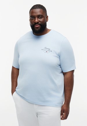 Mann mit Bart, der ein hellblaues Tommy Hilfiger T-Shirt und weiße Hosen trägt, steht mit einer Hand in der Tasche vor einem einfarbigen Hintergrund.