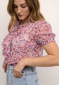 Blusa floreale nei toni di rosa, rosso e blu con dettagli arricciati sul collo e maniche a sbuffo, abbinata a pantaloni shorts in denim azzurro.