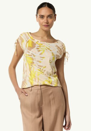 Short-sleeve, light fabric top with abstract yellow and beige pattern, featuring tied accents at shoulders. Worn with beige trousers.