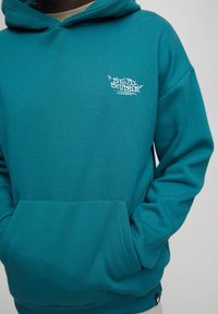 Persona che indossa una felpa color teal con tasca a marsupio e logo bianco "STWD STUDIOS" sul lato sinistro del petto, mani infilate nella tasca.