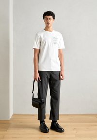 Bílý bavlněný tričko s zeleným potiskem, spárované s šedými pruhovanými kalhotami. Model drží černou crossbody tašku a má na sobě černé boty.
