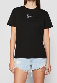 T-shirt en coton noir à manches courtes et col rond, avec une broderie signature blanche sur la poitrine, associé à un short en denim déchiré.