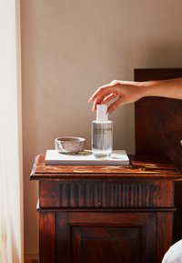 Klare Glas-Haussprühflasche mit weißem Deckel, neben einer runden, marmorierten Keramikschale auf einem hölzernen Nachttisch mit strukturierter Oberfläche.