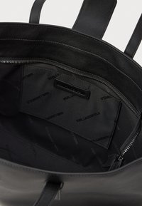 Melnas ādas rokassomas iekšpuse ar rāv�ējslēdzēja kabatu un ar zīmolu apdrukātu auduma oderi; tam ir divas īsas rokturi un strukturēta forma.