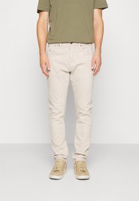 Jeans de mezclilla beige claro con un ajuste slim, diseño de cinco bolsillos y costuras mínimas. Combinados con zapatillas color beige para un look casual.