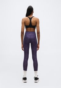 Femme debout de dos portant un soutien-gorge de sport noir, un legging taille haute violet, des chaussettes blanches et des chaussures de sport noires sur un fond uni.