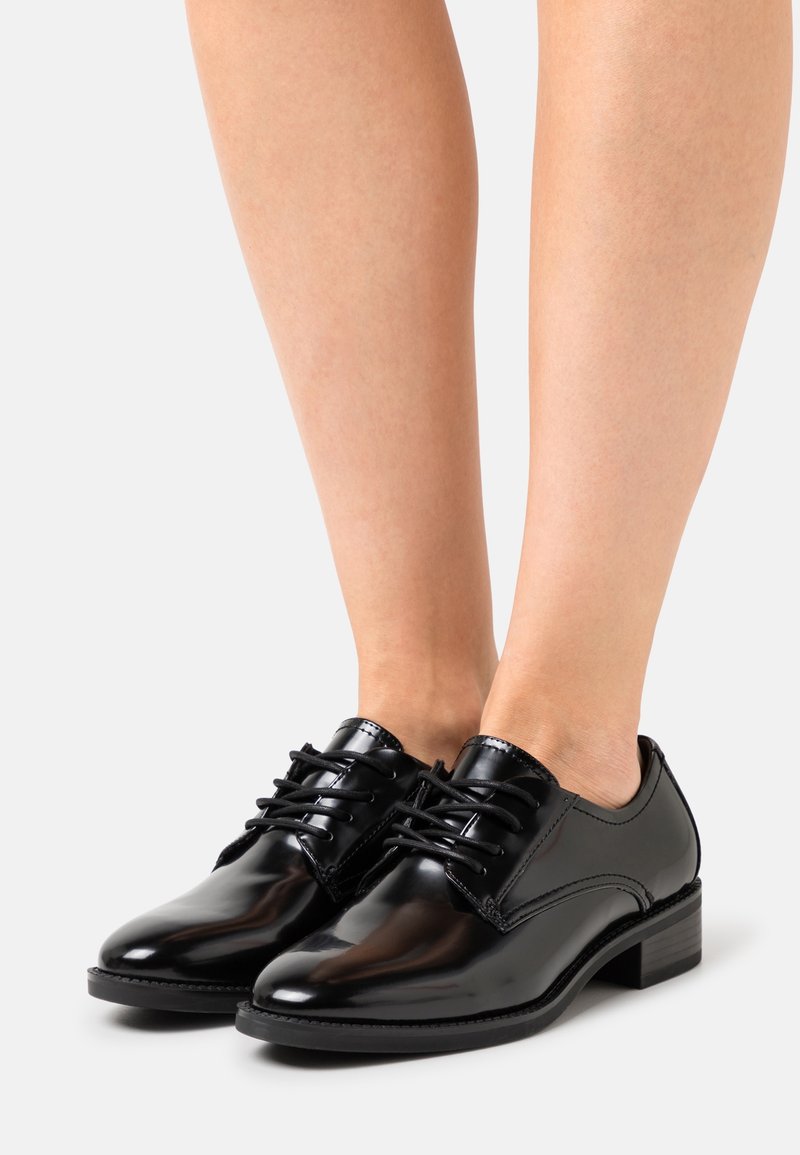 Chaussures Oxford en cuir noir avec finition brillante, design à lacets et petit talon carré. Elles présentent un bout arrondi et des détails de couture.