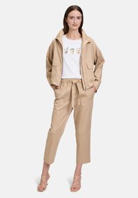Veste cropped beige et pantalon en tissu léger. T-shirt blanc avec des illustrations de boissons à base de café. Sandales beiges à lanières.