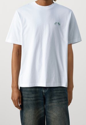 Person iført en ensfarvet hvid kortærmet t-shirt med to små broderede dyr på øverste venstre bryst, kombineret med mørke denimjeans.