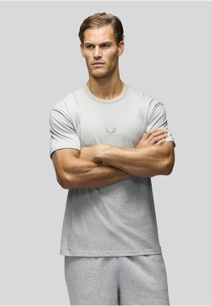 Homme les bras croisés portant un t-shirt gris clair et un short, debout devant un fond gris uni.