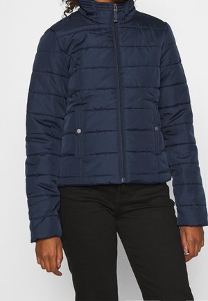 Marineblaue, gesteppte Jacke mit Frontreißverschluss, Taschen mit Druckknöpfen und Stehkragen, getragen zu schwarzer Hose.