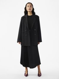 Blazer preto de duas fileiras de botões com lapelas recortadas sobre uma saia pregueada preta a condizer. Sapatos de salto pontudo em bordeaux brilhante. Textura suave.
