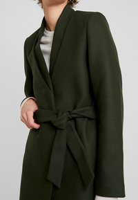 Cappotto avvolgente verde, con texture e cintura a fiocco. Presenta un colletto a scialle e maniche lunghe, indossato sopra un top di colore chiaro.