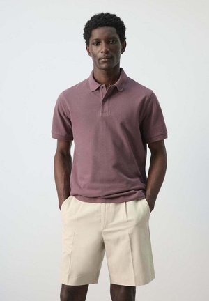 Man with short curly hair wearing a mauve polo shirt and beige shorts, standing with hands in pockets against a plain background.