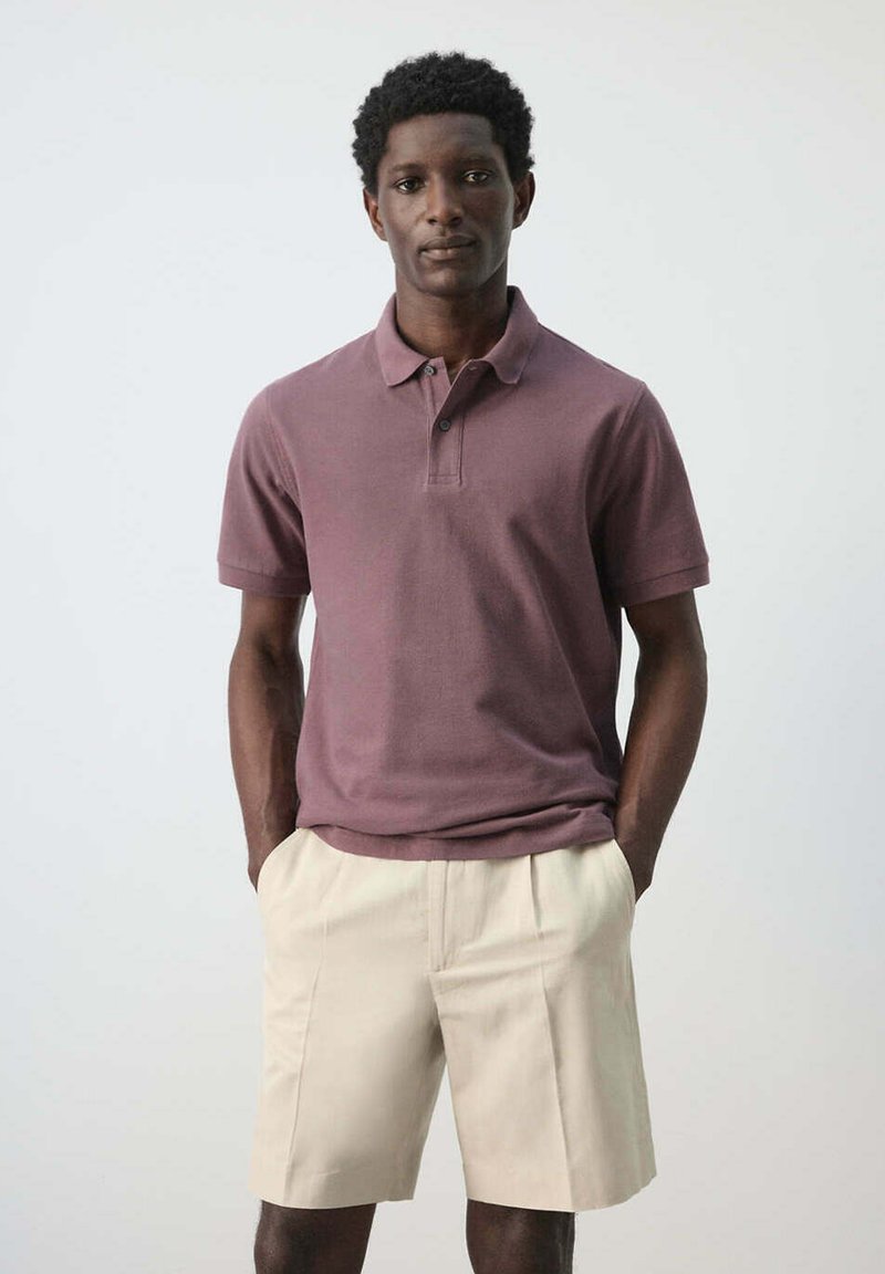 Homme aux cheveux courts et bouclés portant un polo mauve et un short beige, debout les mains dans les poches devant un fond uni.