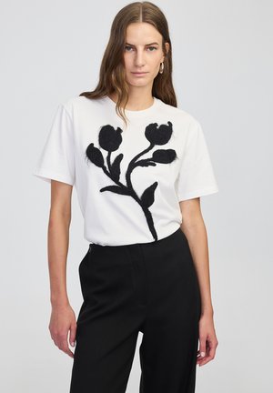 Frau mit langen braunen Haaren, die ein weißes T-Shirt mit schwarzem Blumenmuster und schwarze Hosen trägt, steht vor einem einfarbigen hellen Hintergrund.