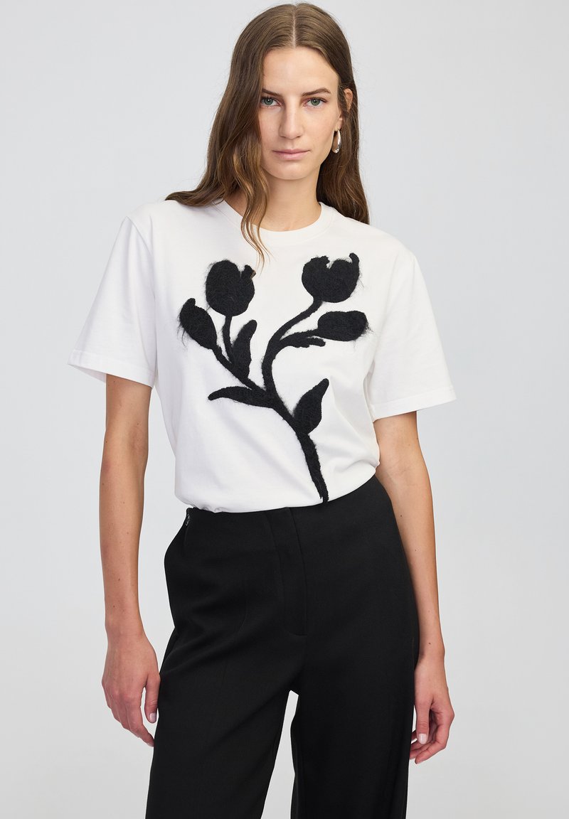 Vrouw met lang bruin haar, gekleed in een witte T-shirt met zwart bloemenontwerp en zwarte broek, staand tegen een effen lichte achtergrond.