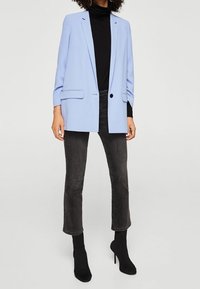 Blazer azzurro chiaro con design su misura, due tasche frontali e chiusura con un solo bottone, indossato sopra un dolcevita nero e jeans grigi a vita alta.