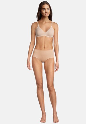 Giovane donna in piedi a piedi nudi, indossa un reggiseno beige senza cuciture e slip a vita alta su uno sfondo chiaro uniforme, guarda leggermente di lato.