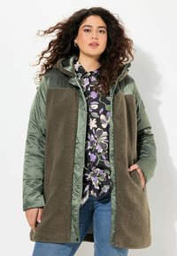 Grön och brun huva coat med ett kviltat mönster, som har en ludig, texturerad front och sidofickor. Bärs över en blommig skjorta.