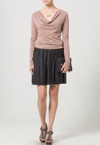 Long-sleeve, blush top with cowl neck and gold polka dots, paired with a black pleated skirt. Accessory includes a black handbag and gray sandals.