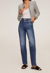 Donna che indossa jeans blu a vita alta, top bianco senza maniche, giacca corta con motivo e sandali neri piatti intrecciati, in piedi di fronte a uno sfondo neutro.