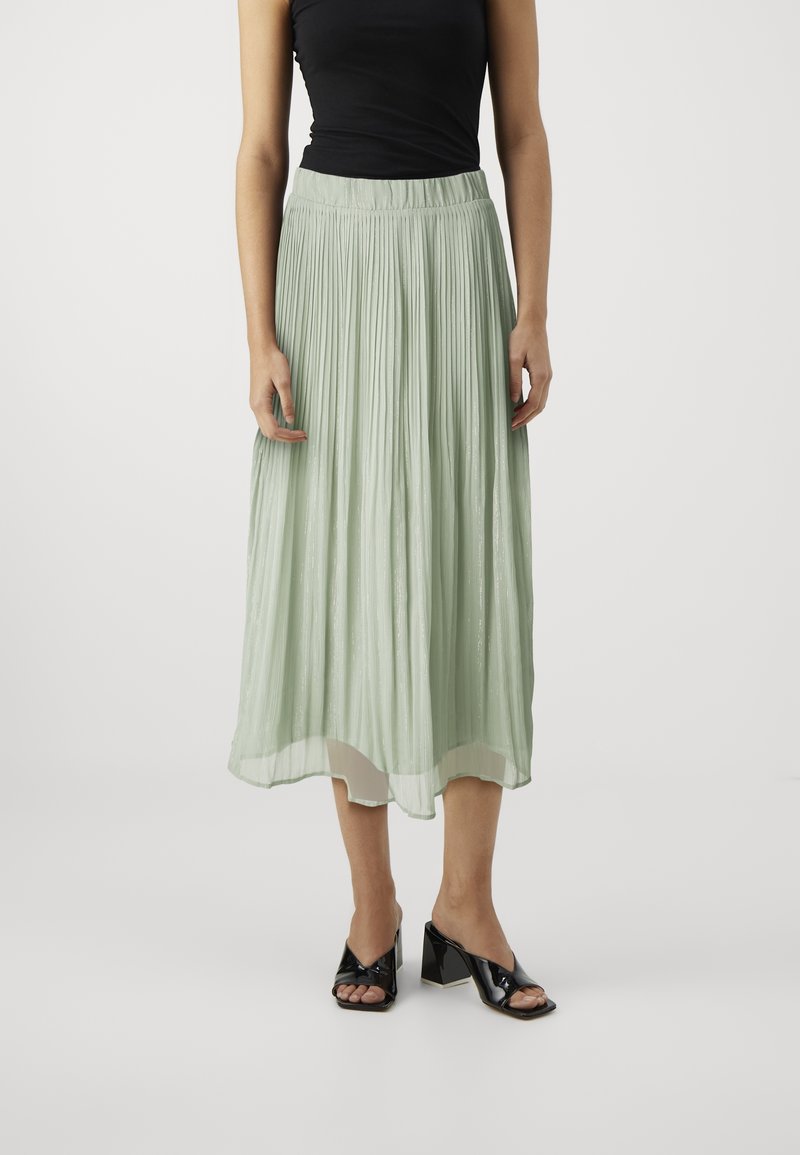 Femme portant une jupe midi plissée vert clair, un haut sans manches noir et des chaussures à talons carrés noires à bout ouvert, debout devant un fond uni.