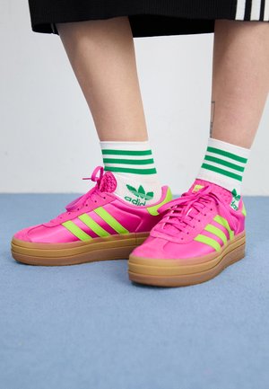 Felle roze sneakers met groene strepen, rubberen zool en textuur bovenkant. Draagt met witte sokken met groene strepen en logo.