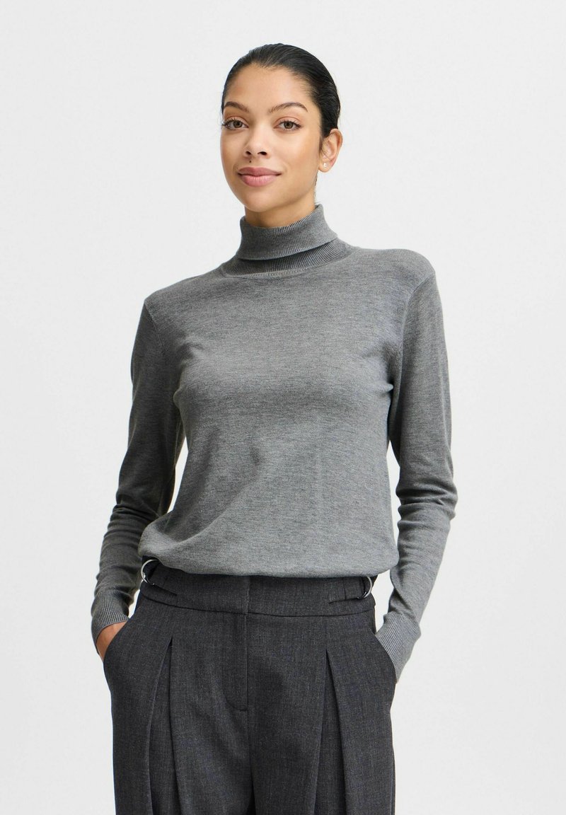 Un pull à col roulé gris en tissu doux avec des manches longues et des poignets côtelés, associé à un pantalon plissé sombre taille haute.